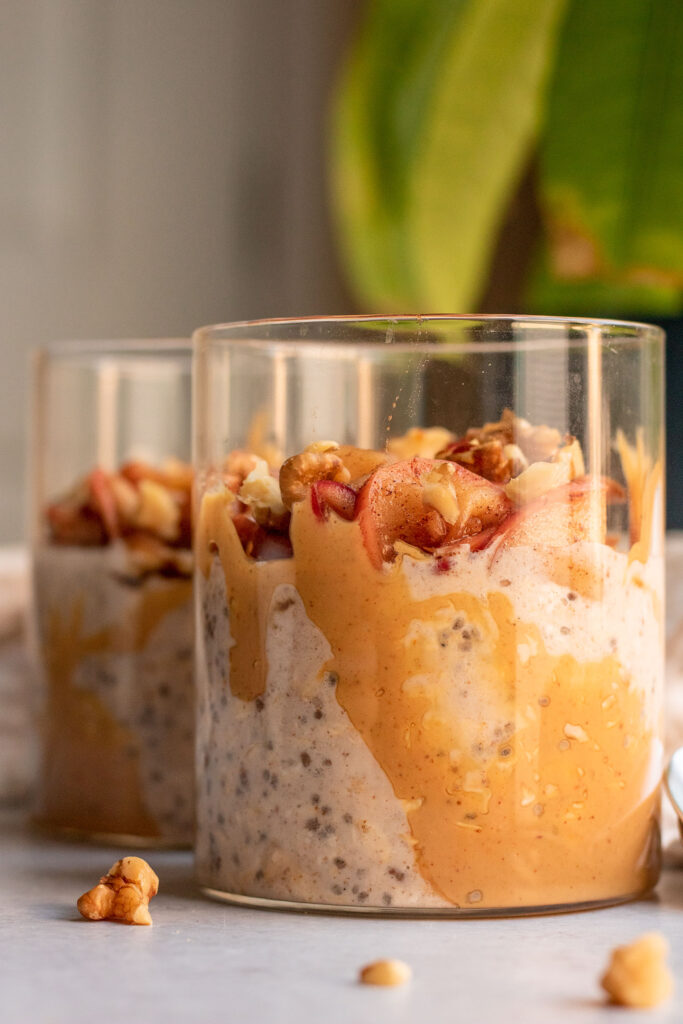 Side view of a peanut butter smeared jar filled with overnight oats and topped with sauteed apples and walnut pieces.