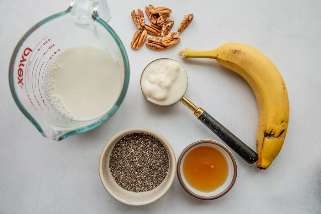 Banana, milk, yogurt, pecans, maple syrup and chia seeds on a working surface.