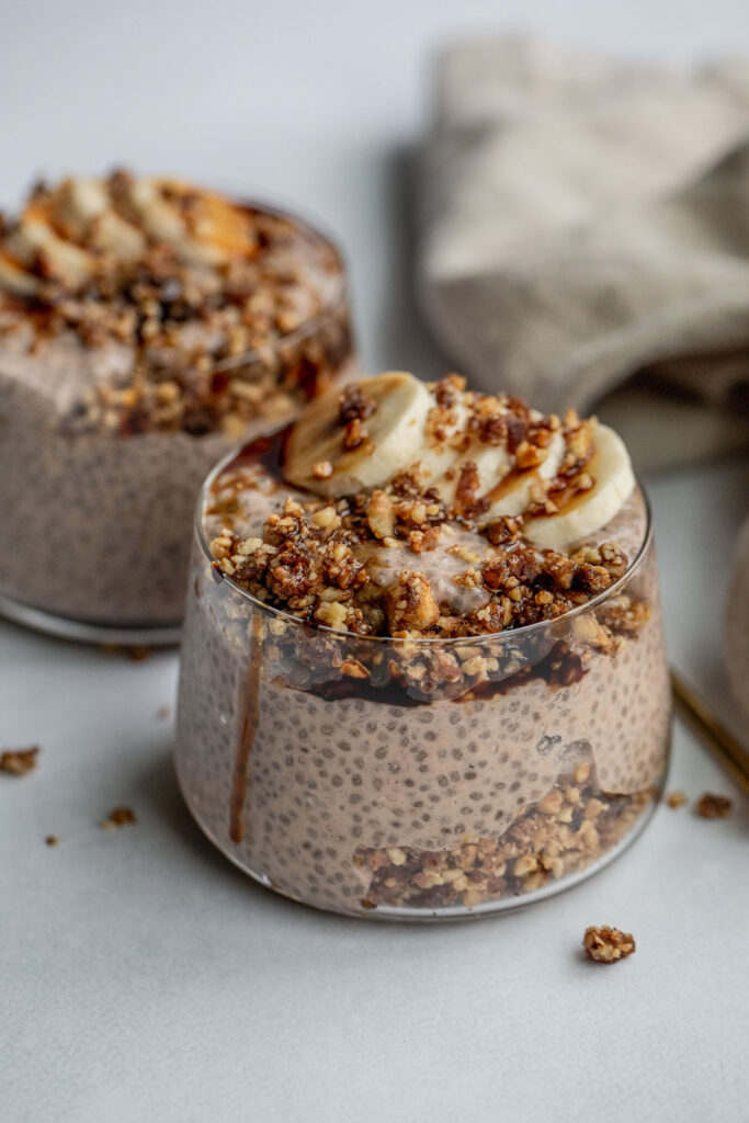 Two glass jars layered with banana chia pudding, pecan date crumble, banana slices and date syrup.