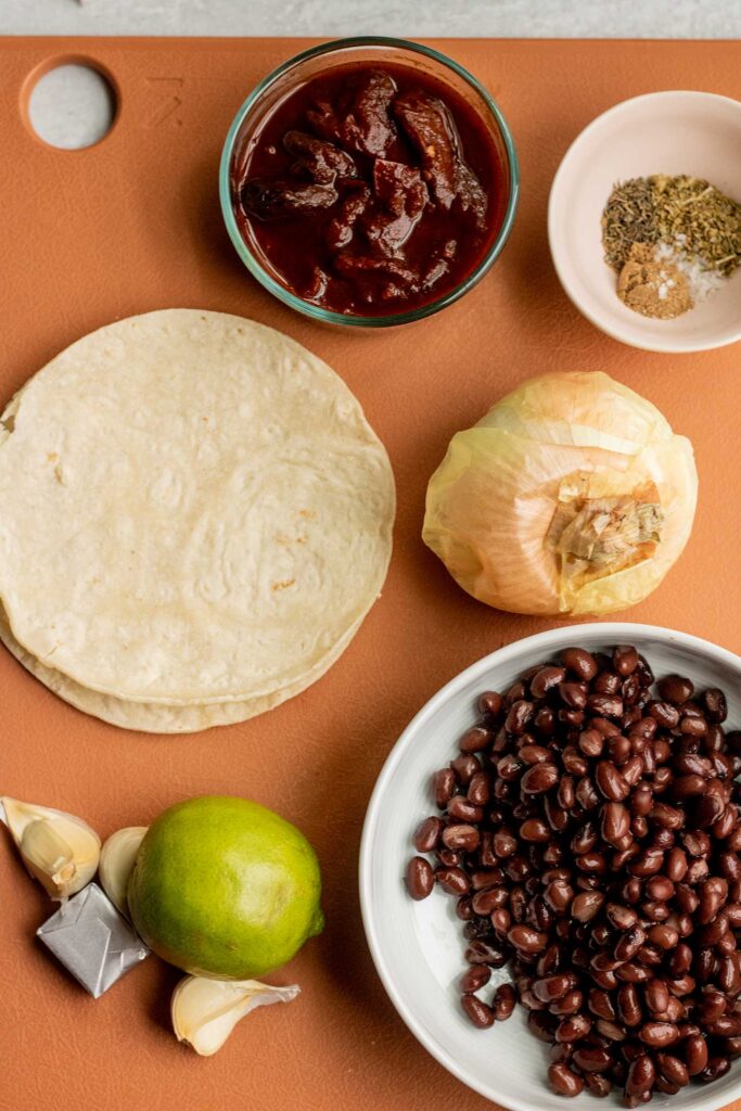 Cutting board topped with onion, black beans, garlic, lime, chipotle peppers and a bowl of seasonings.