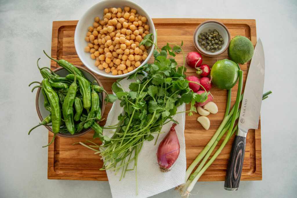 Cutting board topped with a bowl of chickpeas, a bunch of cilantro, shishito peppers, shallot, scallions, garlic cloves, radishes, limes, and capers.