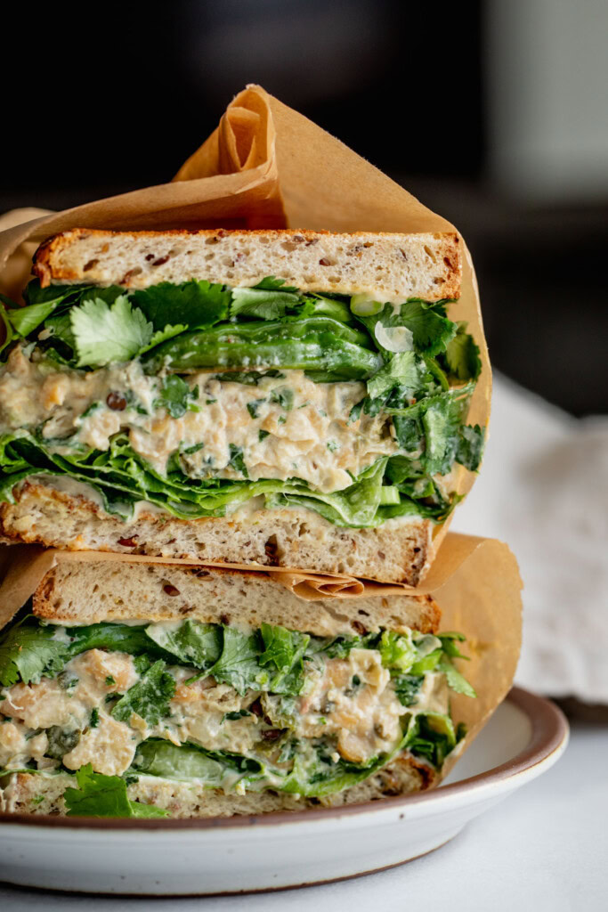 Side view of a sandwich cut in half and stacked on top of itself showing layers of tahini sauce, lettuce, chickpea salad, shishito peppers, cilantro, and scallions between toasted bread.
