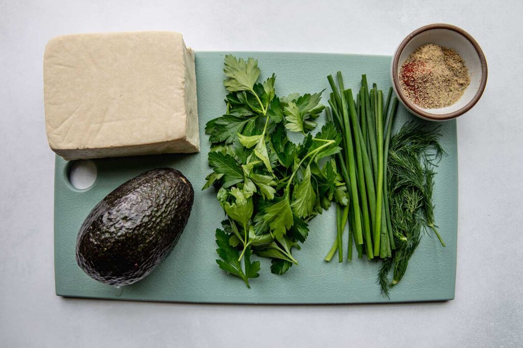 Cutting board with herbs, tofu, avocado and a bowl of seasonings.