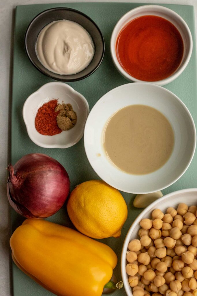 Cutting board with tahini, buffalo sauce, chickpea, bell pepper, lemon, onion, seasoning and yogurt.