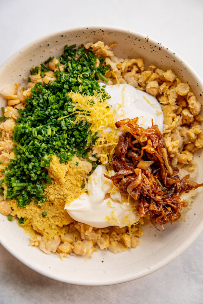 Mixing bowl filled with mashed chickpeas, mayo, yogurt, caramelized onions, nutritional yeast and chives.