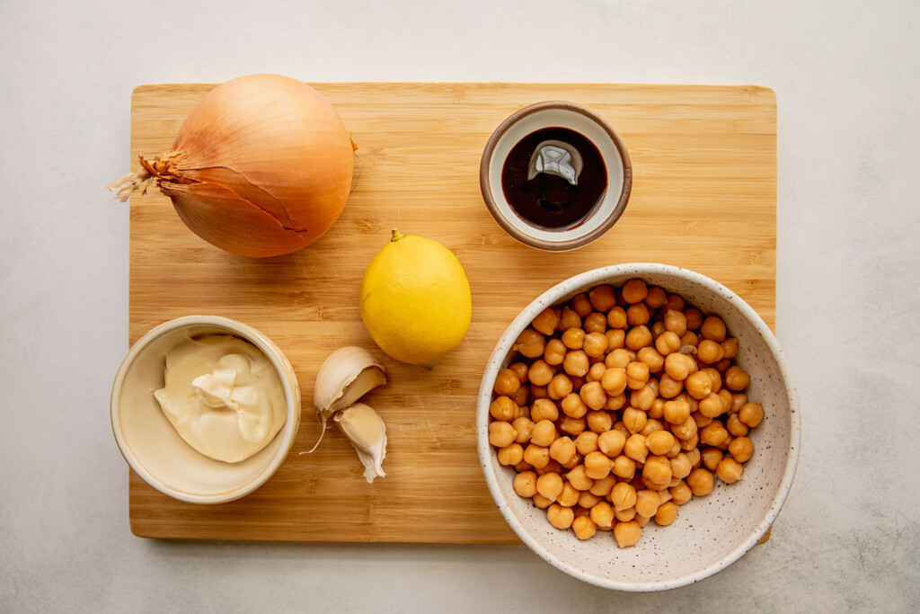 Cutting board topped with onion, lemon, balsamic vinegar, mayo, garlic and a bowl of chickpeas.