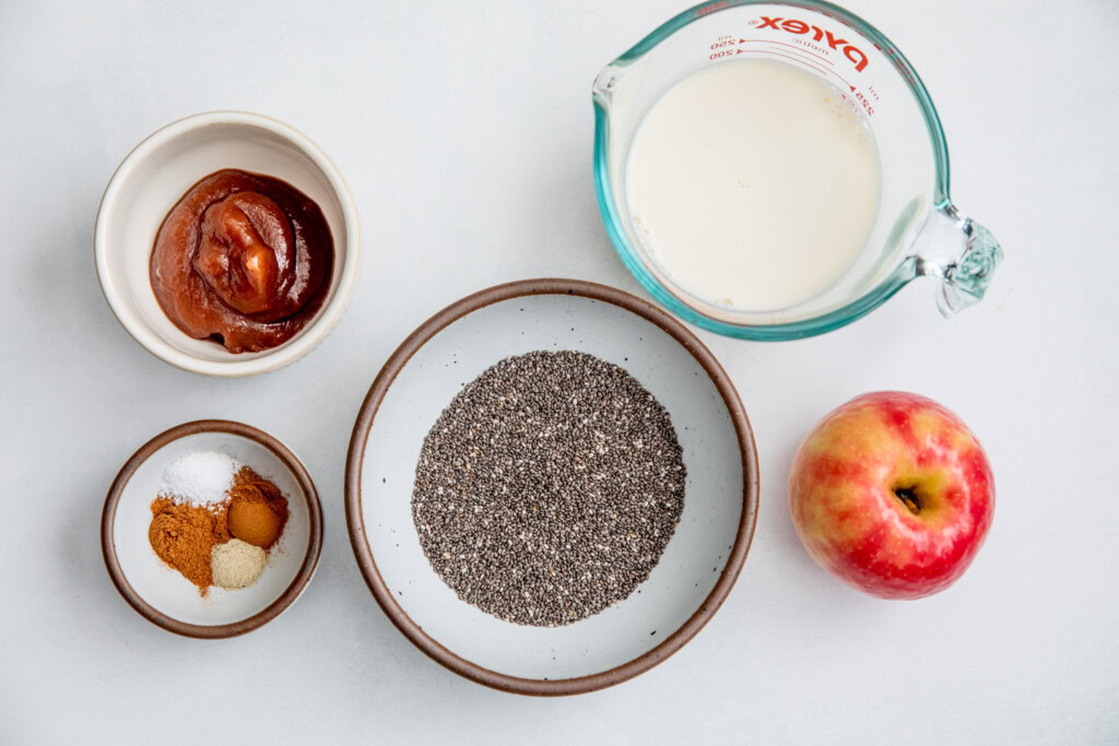 Bowl of chia seeds, cup of milk, apple, bowl of spices and bowl of apple butter placed on a working surface.