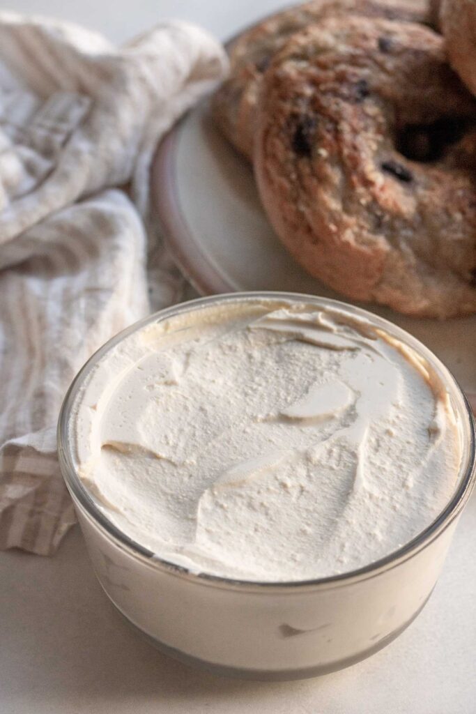 A container of tofu cream cheese in front of a plate of blueberry bagels.