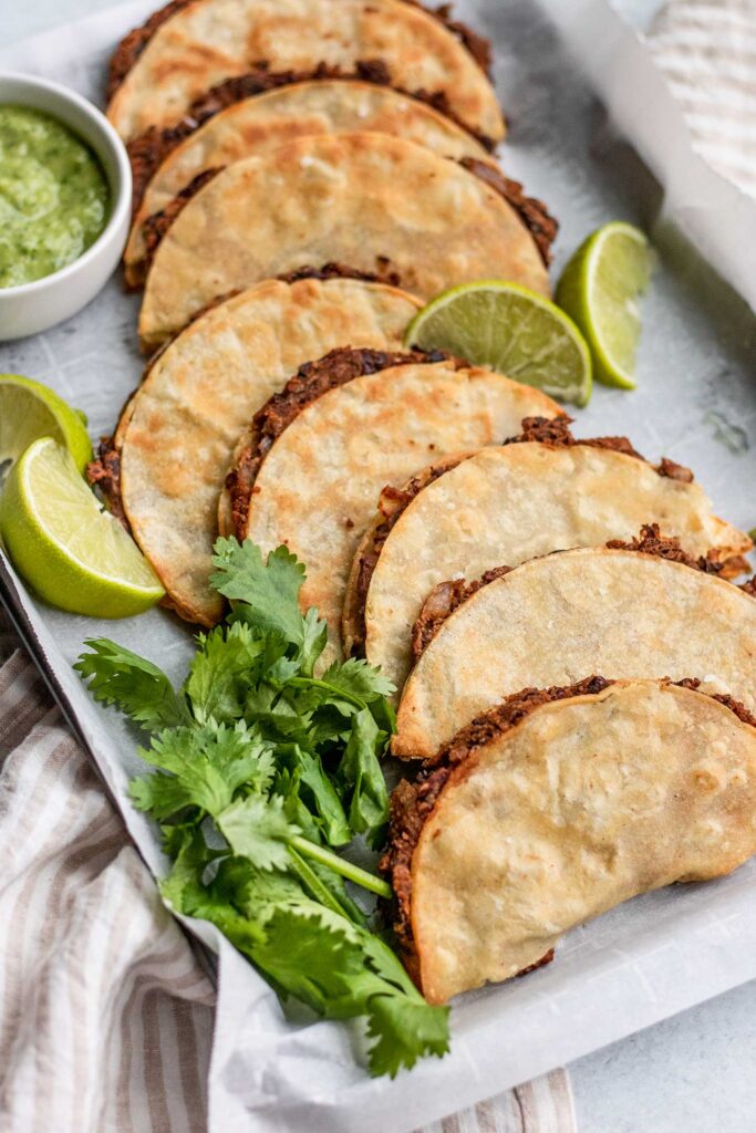A row of black bean tacos arranged on a baking tray lined with parchment paper and topped with lime wedges and cilantro.