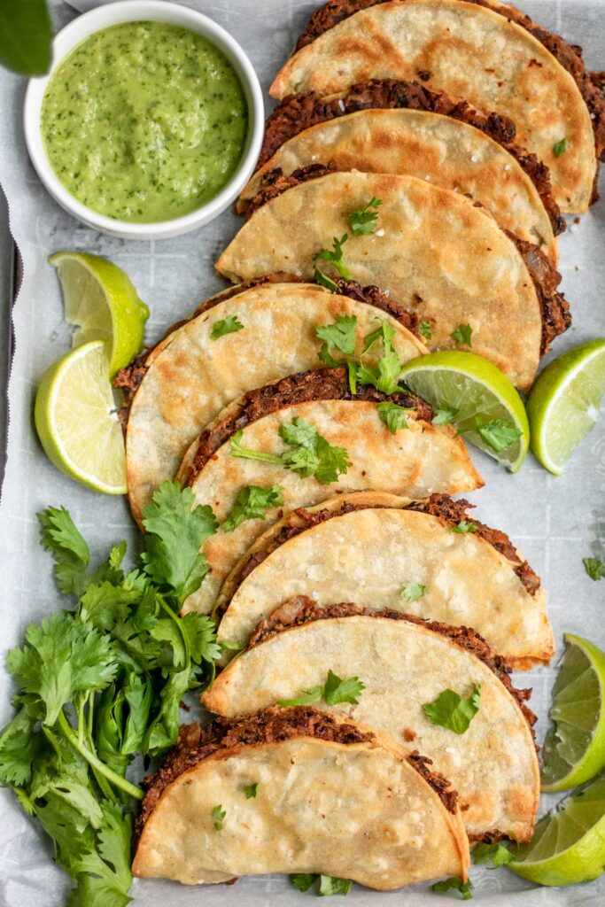 Top down view of a row of tacos arranged on a baking tray with lime wedges, cilantro lime sauce and cilantro.