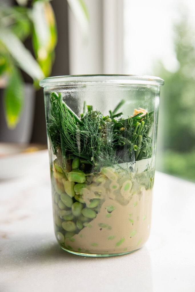 Glass container filled with edamame, tahini, garlic, chives, dill and oil.