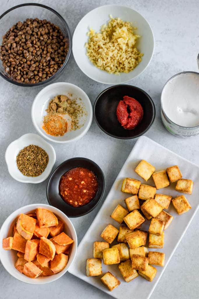 Tofu, roasted sweet potato, ginger, garlic, tomato paste, garlic chili sauce, spices, coconut milk, and lentils set up ready to cook.
