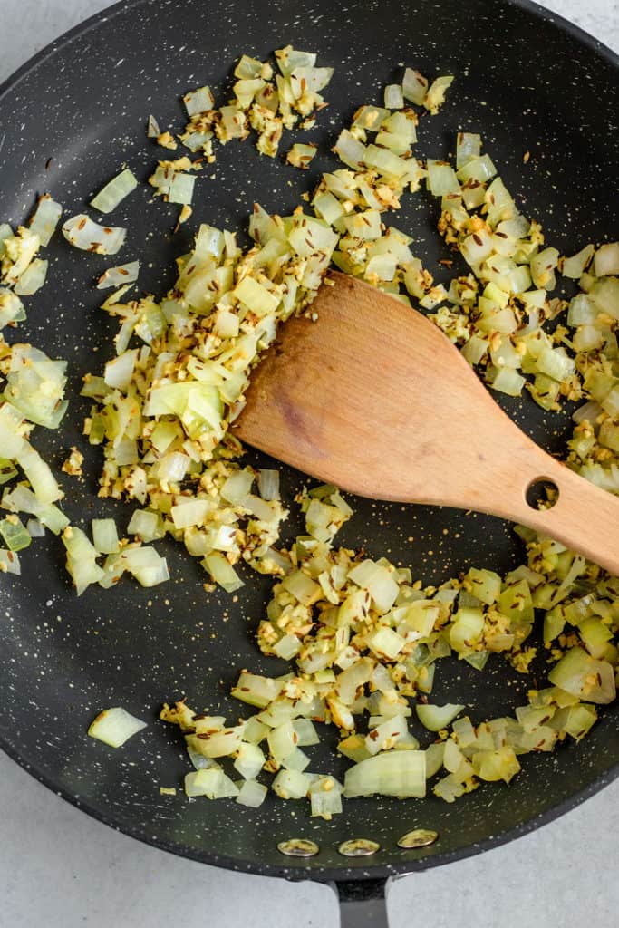 Onions, aromatics and spices sauteing together in a large black pan.