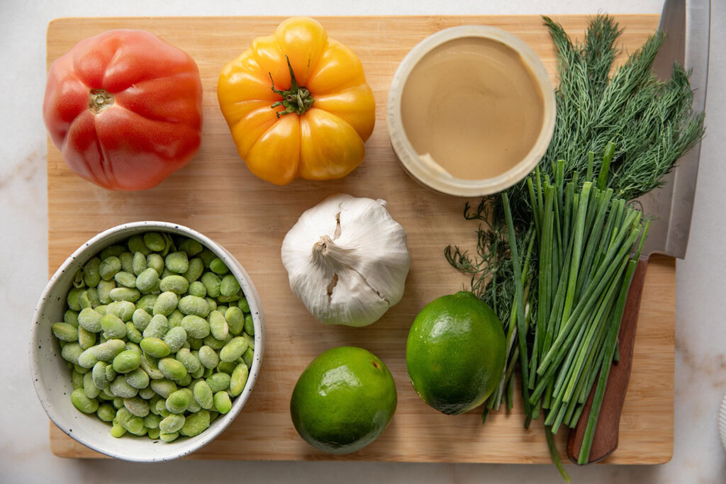 Cutting board topped with heirloom tomatoes, bowl of edamame, limes, chives, dill, garlic and tahini.