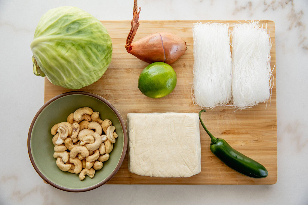 Cutting board with tofu, jalapeno, cashews, mung bean noodles, shallot, lime, and cabbage.
