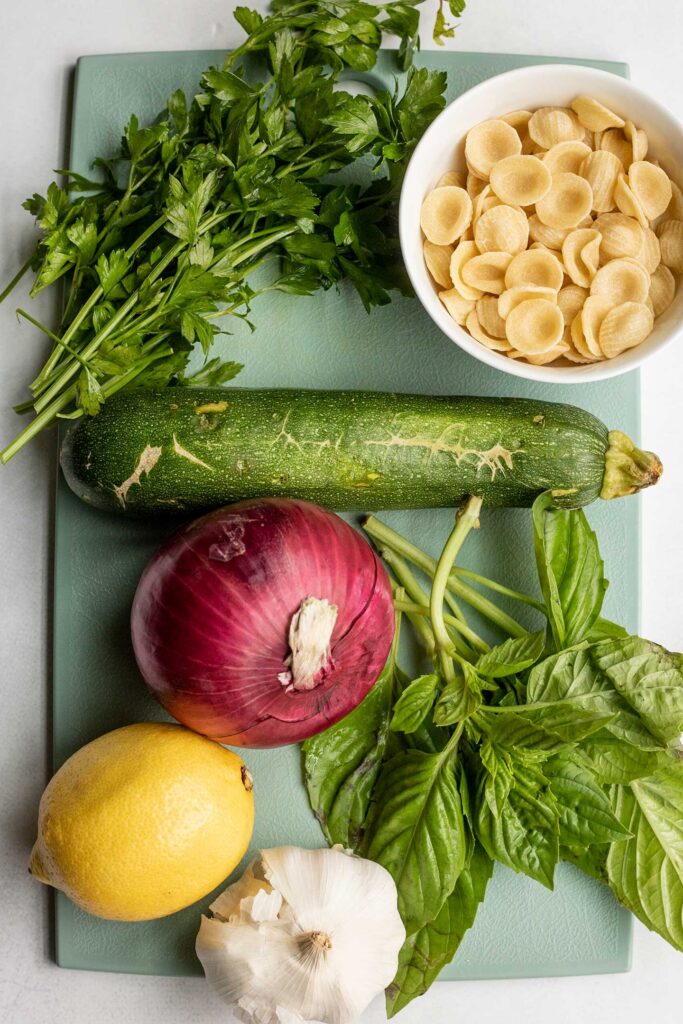 Cutting board with zucchini, onion, lemon, basil, parsley and pasta on top.