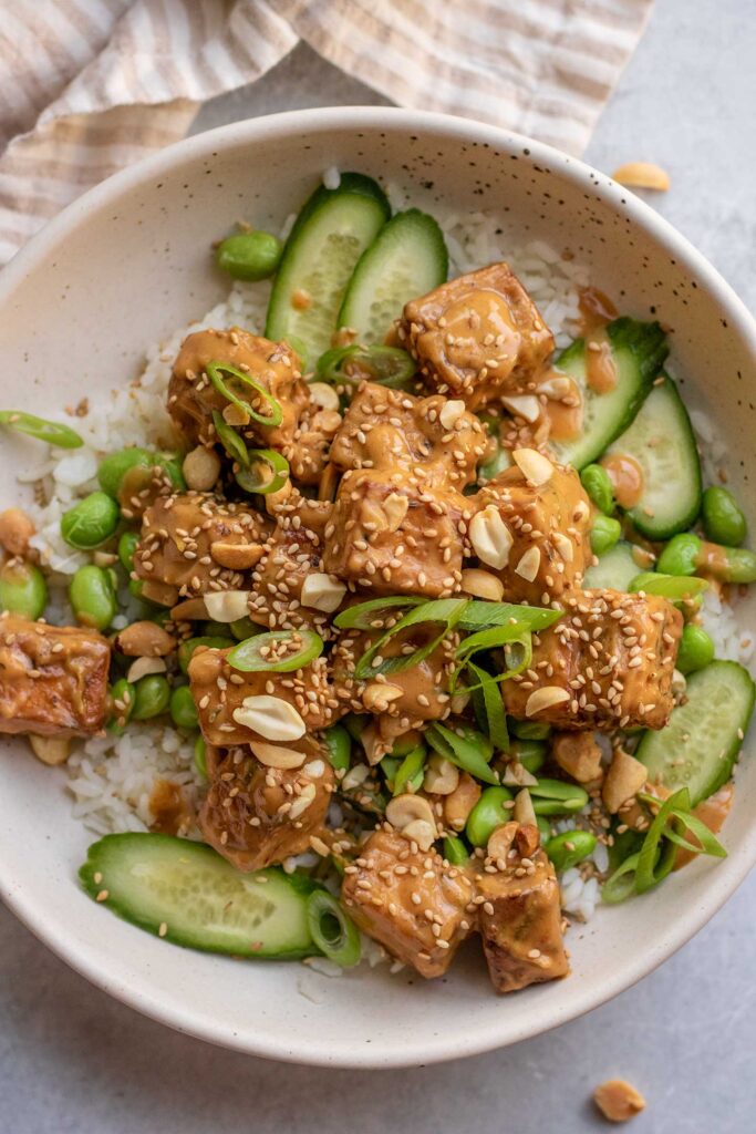 Top down view of a bowl of rice, edamame and cucumbers topped with baked tofu coated in a peanut sauce.