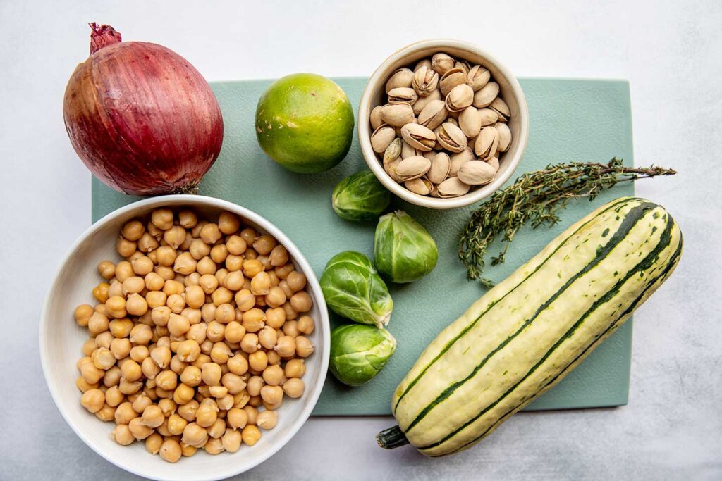 Cutting board with an onion, lime, brussels sprouts, thyme, delicata squash, and chickpeas.