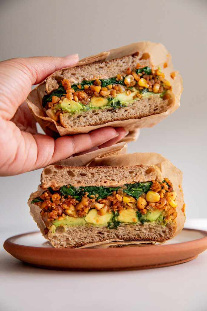 Picking up half of a cut breakfast sandwich that shows it is stuffed with tempeh hash, avocado and spinach.