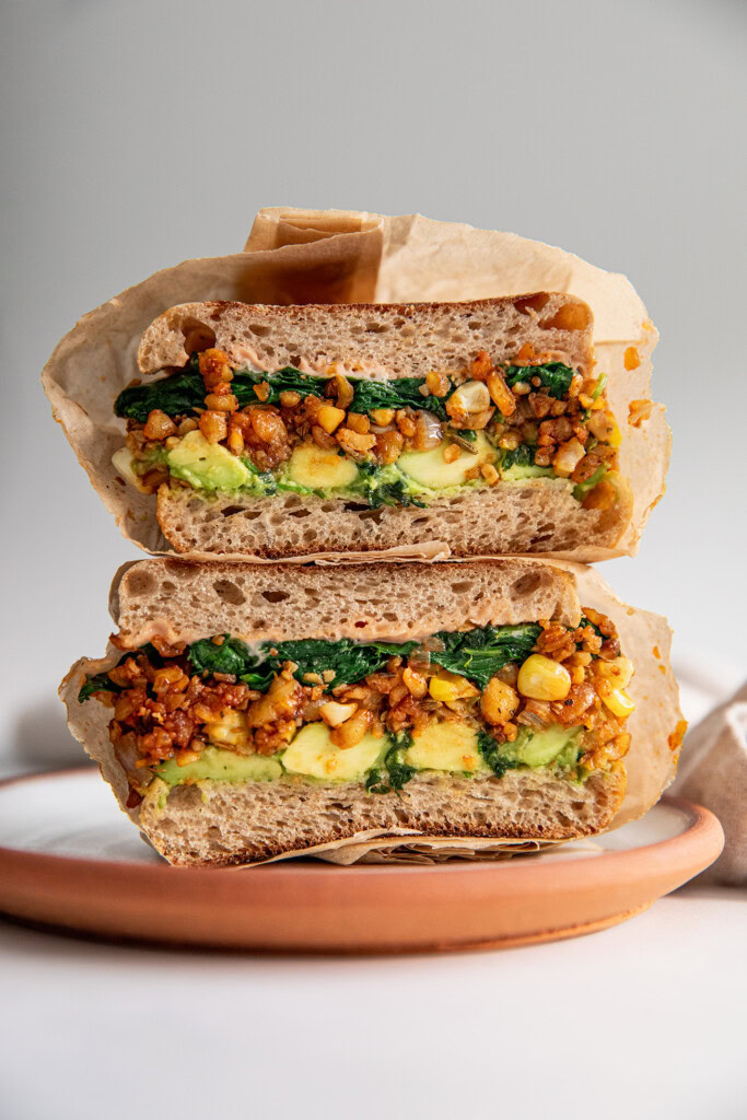 Breakfast sandwich stacked on a plate and stuffed with mashed avocado, smoky tempeh hash, and spinach.