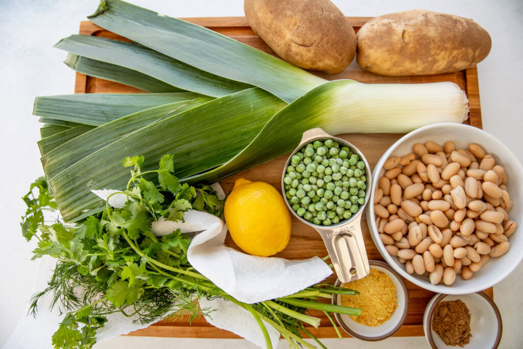 Cutting board topped with a leek, potatoes, lemon, peas, white beans, nutritional yeast, and herbs.