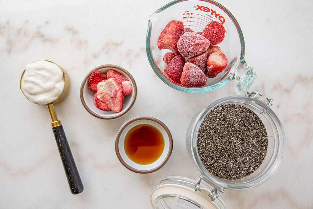 A working bench topped with a cup of frozen strawberries, chia seeds, maple syrup, yogurt, and freeze dried strawberries.