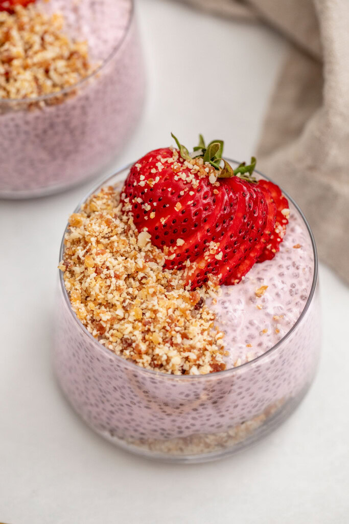 A close up of a layered cup of almond date crumble and strawberry chia pudding then topped with a sliced strawberry on top.
