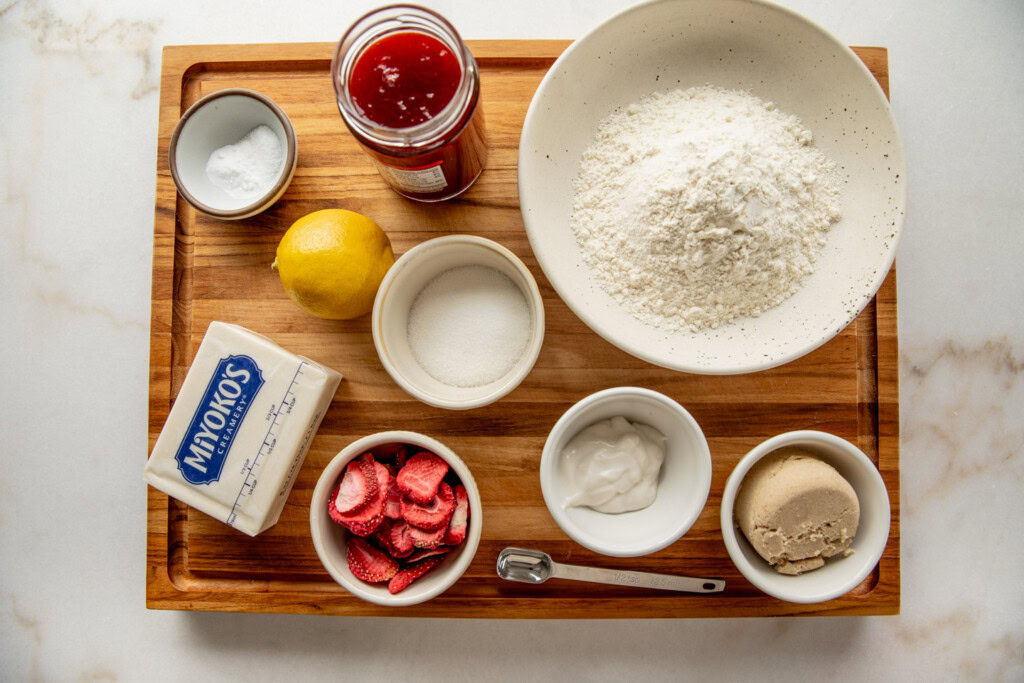 Cutting board with a bowl of flour, freeze-dried strawberries, brown sugar, white sugar, lemon, baking soda, and vegan butter.