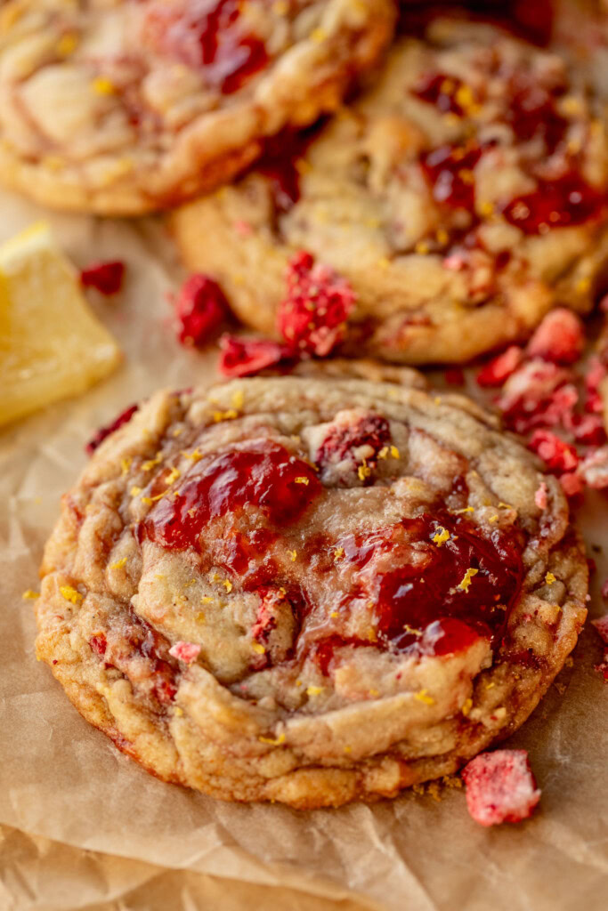 Close up of a strawberry jam cookie surrounded by other cookies, lemon slices and crushed freeze-dried strawberries.