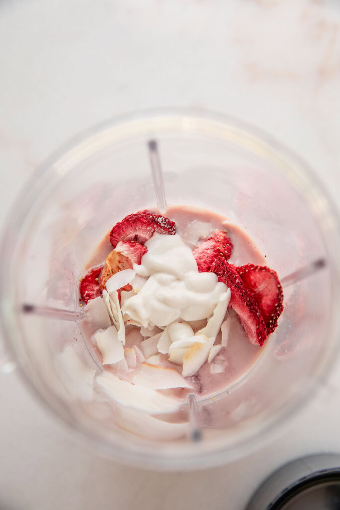 Blender cup filled with strawberries, milk, yogurt, and coconut.