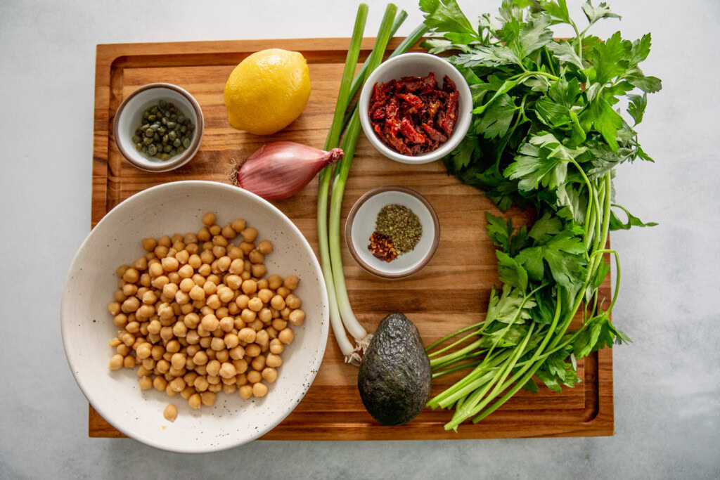 Cutting board topped with a bowl of beans, avocado, parsley, scallions, oregano, pepper, shallots, lemon, capers, and sun-dried tomatoes.