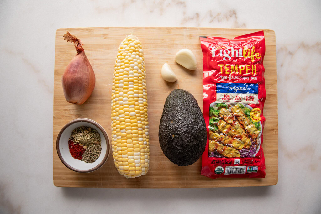 Cutting board topped with a block of tempeh, avocado, corn, garlic, spices and shallot.
