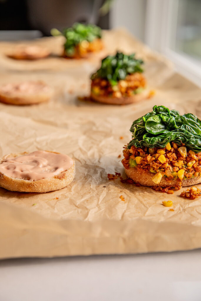 Assembling breakfast sandwiches all in a row topped with avocado, tempeh hash and spinach.